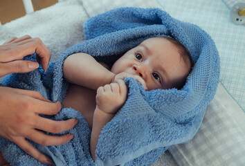 Unrecognizable Mother Wrapping her Clean Baby Boy in a Towel After Bath at Home. 

An anonymous mother hands drying her cute baby with blue towel on a baby bed.