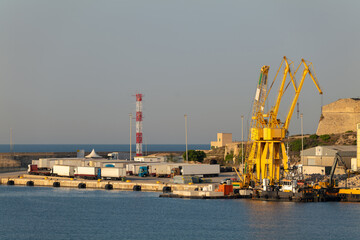 In the morning after sunrise in the port of Palma on the Mediterranean island of Mallorca. There are cranes on the right and a lot of trucks waiting on the left. A working platform is on the water.