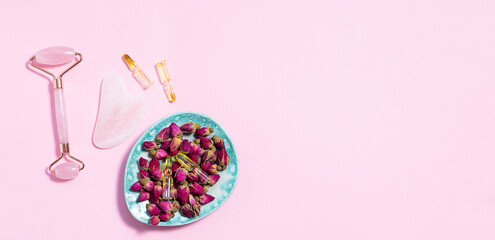 Facial massage equipment, ampules with organic massage oil and dry rose flowers on light pink background, copy space for your design. Skin care concept.