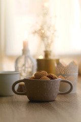 Bowl of cinnamon cookies, cup of warm drink, lit candles and vase with gypsophila flowers on the table. Selective focus, fairy lights in the background. Hygge at home.