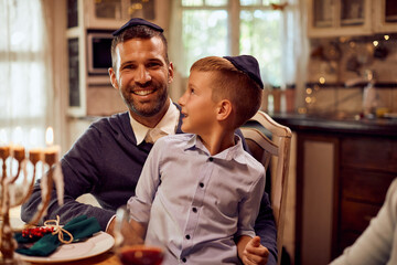 Happy father and son wear yarmulke while celebrating Hanukkah at dining table at home.