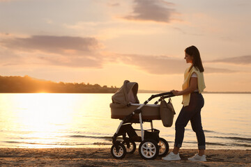 Fototapeta na wymiar Happy mother with baby in stroller walking near river at sunset