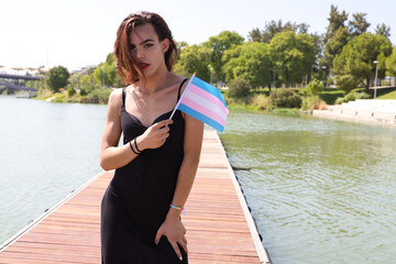 young latina and transgender woman on the boards of a river pier holding the gay pride flag. Concept diversity, transgender, and freedom of homosexual expression.