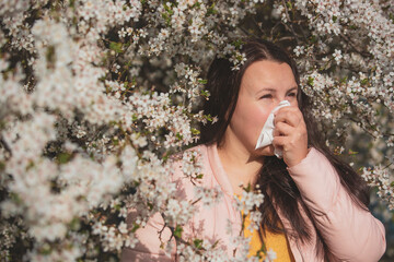 Spring allergy symptoms concept, young woman sneezing in front of blooming a tree, allergy to pollen and flowering season, healthcare