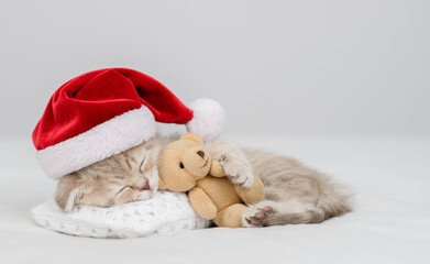Cute kitten wearing red santa's hat sleeps under white blanket and hugs favorite toy bear