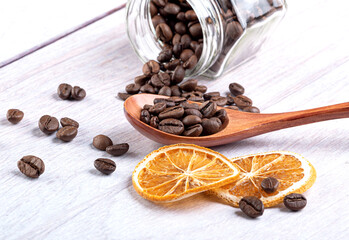 Coffee beans in a wooden spoon, spilling out of a glass jar, dried oranges on a light wooden background.