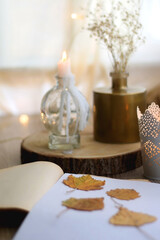 Old book with pressed autumn leaves, lit candles and vase with gypsophila flowers. Selective focus.