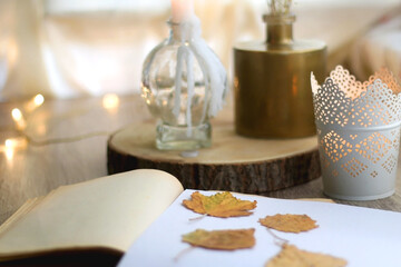 Old book with pressed autumn leaves, lit candles and vase with gypsophila flowers. Selective focus.