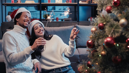 smiling african american couple holding sparklers near christmas tree