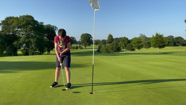 Young boy on a golf green putting a golf ball into the hole and celebrating afterwards
