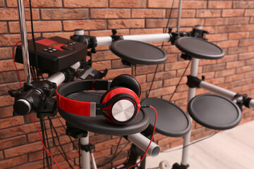 Modern electronic drum kit with headphones near red brick wall indoors. Musical instrument