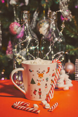 Obraz na płótnie Canvas mug with marshmallows and striped caramel on a red background against the background of an elegant Christmas tree