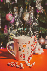 mug with marshmallows and striped caramel on a red background against the background of an elegant Christmas tree