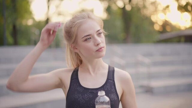 Sport and Fitness. Young Caucasian Woman Dressed Sportswear Standing Outdoor Drink Water Plastic Bottle Summer Park Sunlight Background Resting After Training Jogging Running Exercises loosens hair