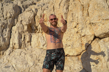 Young tattooed sporty man on the beach on a hot summer sunny day against the backdrop of a mountain posing in sunglasses