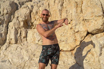 Young tattooed sporty man on the beach on a hot summer sunny day against the backdrop of a mountain posing in sunglasses