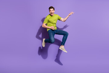 Fototapeta na wymiar Full size photo of cool young brunet guy jump play guitar wear t-shirt jeans sneakers isolated on violet background