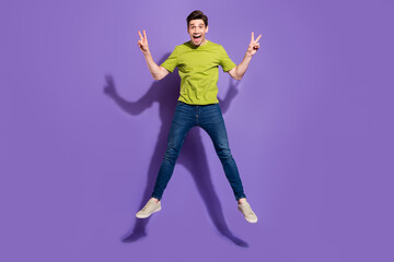 Fototapeta na wymiar Photo of cute charming young man guy wear green t-shirt jumping high showing v-signs smiling isolated violet color background