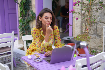 Young woman in a dress in a bright street cafe with a laptop works remotely on her own schedule from anywhere in the world online, freelancer
