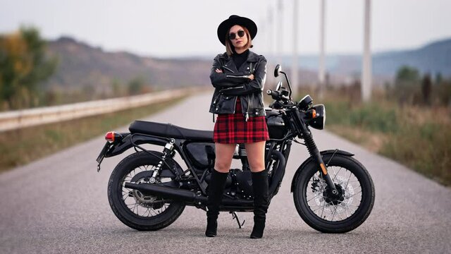 Portrait of motorcyclist woman in mini skirt, leather jacket and hat standing near vintage-styled motorcycle. Driver in jackboots and sunglasses on roadway. Trip, speed, style, feminism concept.