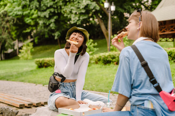 Multiracial two women smiling and eating pizza while resting in park