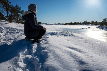 Fototapeta na wymiar Woman in warm clothing and knitted beanie sitting down in the snow, enjoying the sun while looking at the winter landscape in the Swedish archipelago with its frozen water.