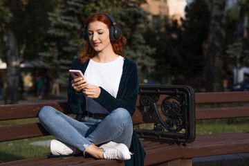 A beautiful, attractive and happy redhead Caucasian girl with headphones listening to music and using a smartphone while sitting on a bench.