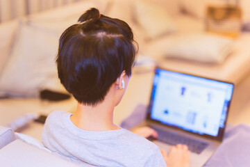 Asian boy with earphone sit on couch working on laptop at home.