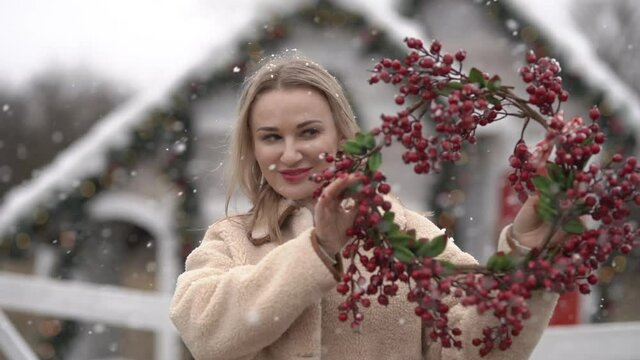 Beautiful blond woman in white winter coat posing with red Christmas wreath against decorated house. Focus is on the wreath. Snowing. Holiday concept. Slow motion.