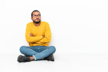 Young Ecuadorian man sitting on the floor isolated on white wall making doubts gesture while lifting the shoulders