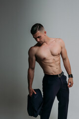 A man with a naked torso in suit trousers. He holds a jacket in hand. The abs muscles, pectoral muscles, large biceps, and a thin waist are clearly visible. Office employee with a bodybuilder's body