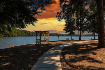 Fototapeta na wymiar a shot of a wooden swing on the banks of the lake near a smooth footpath with still blue lake water surrounded by lush green trees with powerful clouds at Lake Peachtree in Peachtree City Georgia