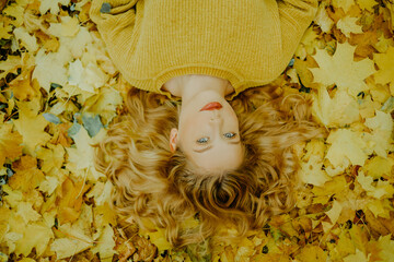 A beautiful young girl lies in the park with autumn leaves, a cute model with beautiful blond hair. Autumn discounts, autumn yellow sweater