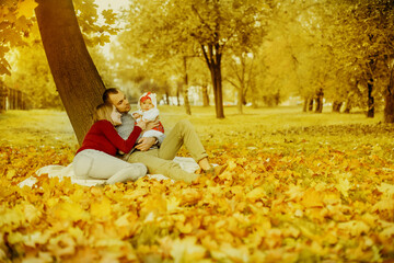 Couple in love sitting on autumn fallen leaves in a park, sit near a tree , enjoying a beautiful autumn day. Happy joyful young family father, mother and little baby  girl having fun outdoors.