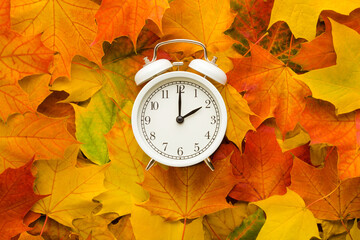 White alarm clock on colorful maple leaves background. Closeup. Time change concept. Top down view.
