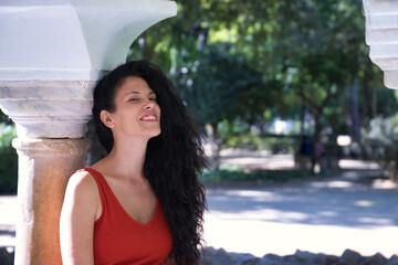 Middle-aged adult Hispanic woman with black curly hair, leaning on a column, smiling. Concept happiness, joy, sensations, looks.