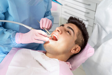 Young man having teeth examination at dentist office. Professional oral checkup in dentistry. Close up of female dentist working with dental tools in opened patient mouth. Stomatology and teeth care