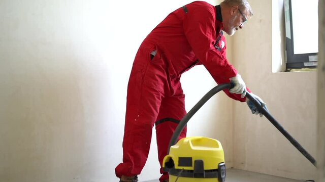 Worker man with orange overalls and glasses hoovering construction works remains and dust with vacuum cleaner in room. Static shot