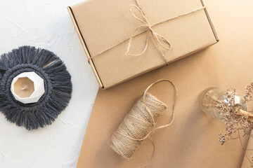 Handmade candle on macrame coaster, present or parcel craft box and jute rope bobbin. Organic packing, eco-friendly parcel. Dry wild herb in vase. White craft paper background, flay lay, top view.