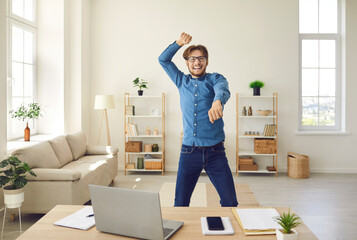 Emotional business man or student happy, excited and euphoric about success, feeling free after finishing work, jumping for joy and dancing gangnam style near desk with laptop computer in home office
