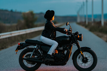 Hipster stylish motorcyclist woman sitting on vintage-styled motorcycle. Young female driver in hat at evening on roadway. Trip, freedom, classic motorbike concept.