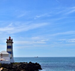 Lighthouse tower on background of blue sky and blue sea with  small ship silhouette on horizon