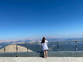 woman in standing near the edge of observation deck viewpoint or balcony with metallic handrails and transparent glass walls looking towards mountains in evening view from back