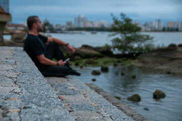 man in blurred image wearing dark clothes sitting on the rock in front of the beach in cloudy afternoon with cell phone in his hands thoughtful, landscape in the background.