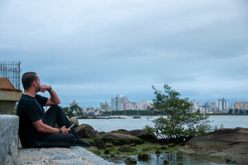 man sitting by the beach on the rock in gray afternoon, wearing dark clothes, thoughtful.