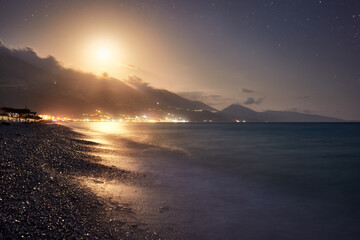 Night view of the beach at Borsh, moon rising over a rock ridge, long exposure, beach lights in the background. Albania travel.