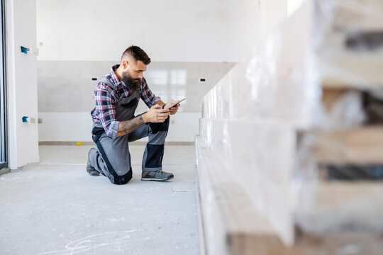 An Innovative Supervisor In Overalls Is Kneeling Inside The New Building And Checking On Materials On His Tablet. He Is Kneeling Next To A Pile Of Beams.