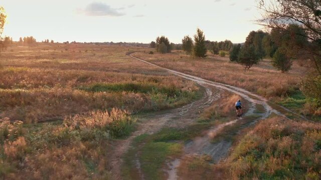 Young caucasian cyclist rides on a gravel bicycle on a countryside road through the field at sunset. Active recreation outdoors. Bicycle sports and travel. Aerial view.