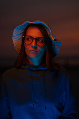 a European woman illuminated by neon light in a hat against the background of the sunset sky. neon lights and a woman at sunset. stylish cyber photo. color filters on neon lamps. creative photo and