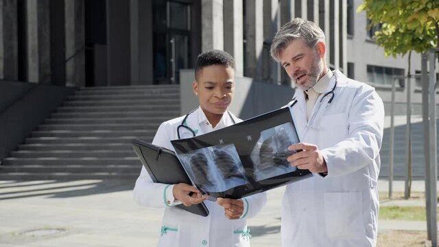 Two professional doctors wearing lab coat stethoscope analyzing X-ray image outdoors stand near hospital main entrance. Internet radiologist exam x-ray lungs during COVID 19 epidemic outside.